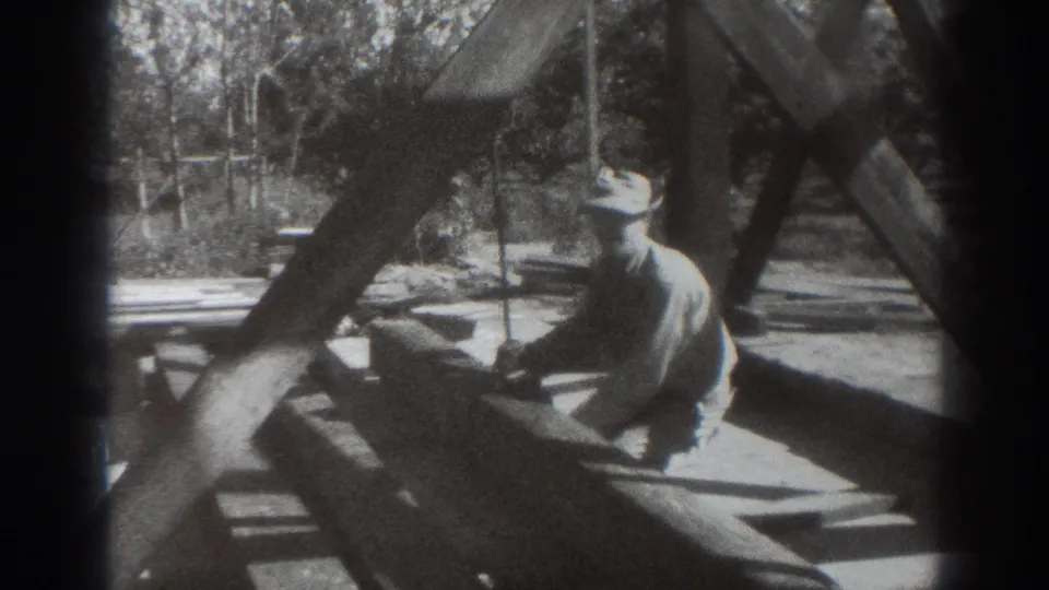 A hat-wearing worker turns while kneeling on or beside a large wooden beam, continuing rope work in the 1937 Sacramento footage.