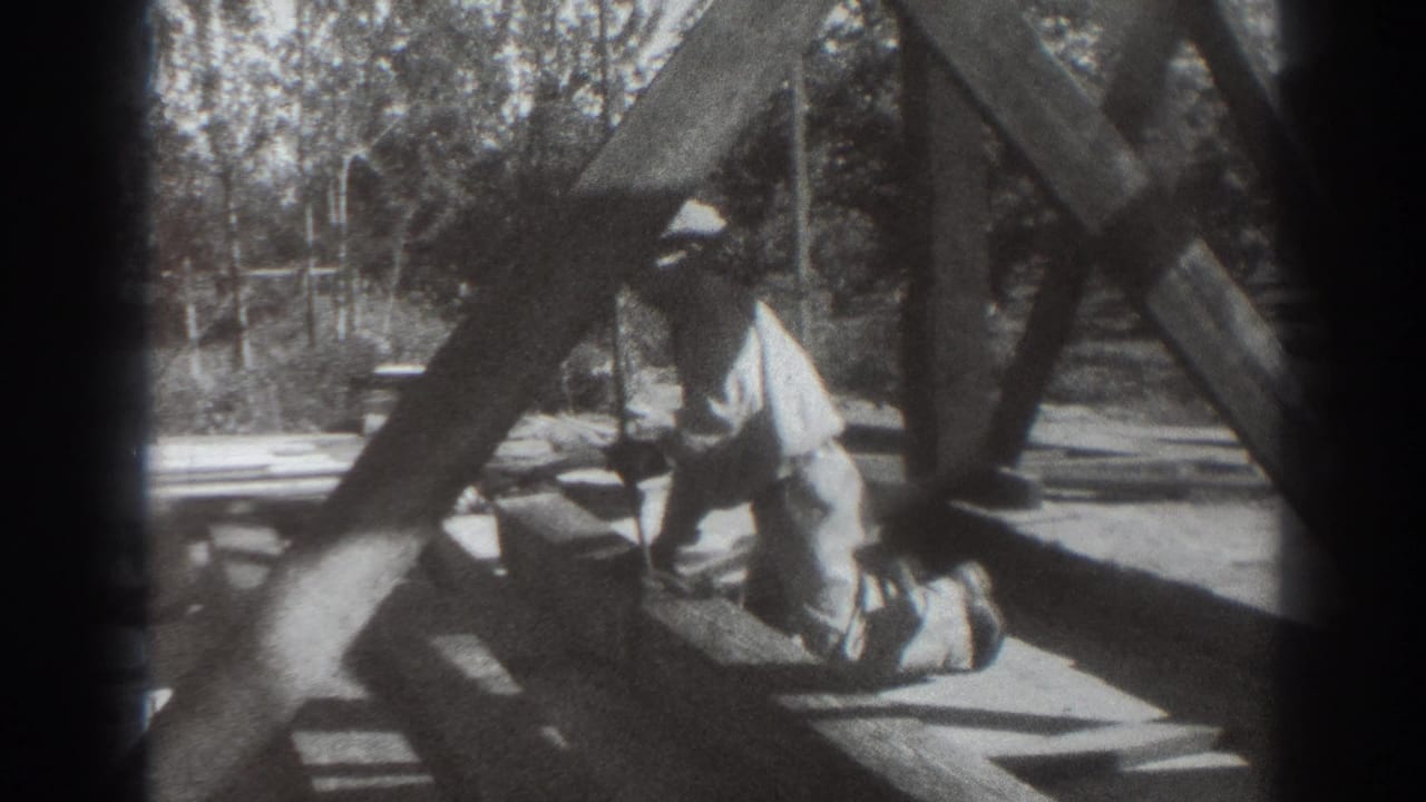 In 1937 Sacramento home-movie footage, a man in a hat kneels at the base of a wooden tower frame with trees behind him.