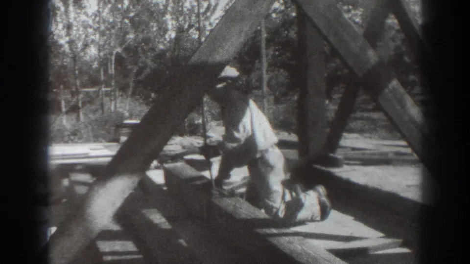 In 1937 Sacramento home-movie footage, a man in a hat kneels at the base of a wooden tower frame with trees behind him.
