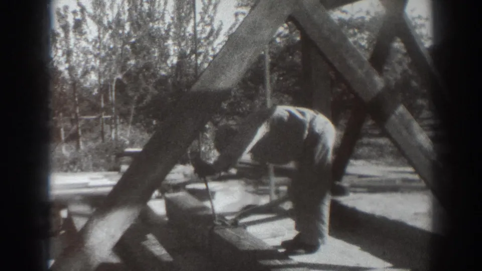 A man in light work clothes bends near the base of the wooden tower structure in Sacramento, with diagonal beams casting shadows.