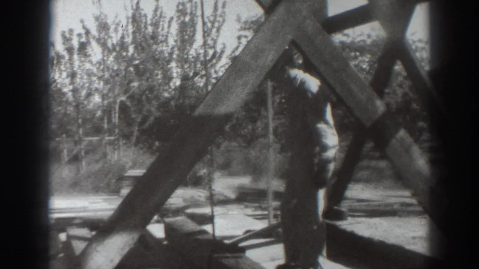 The man stands beside the timber framework, appearing to work with a tool near the base while trees fill the background.