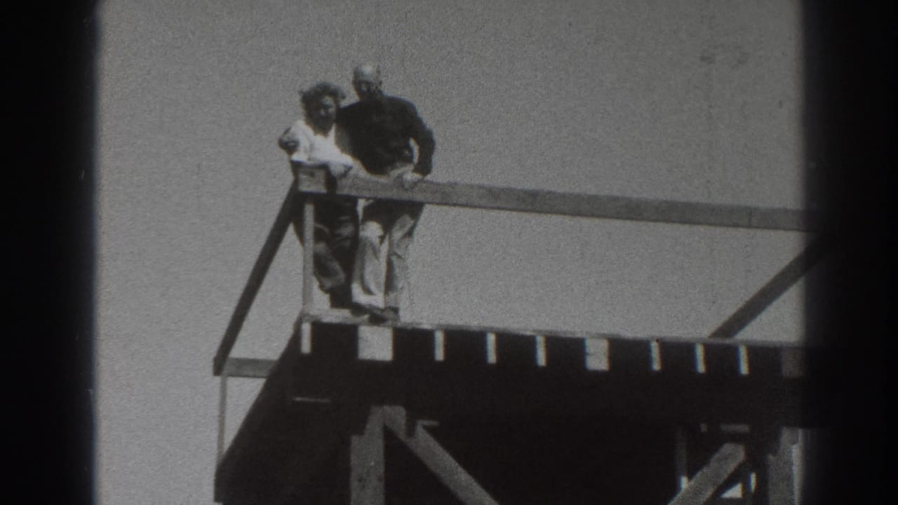 The couple remains at the platform railing, framed by wooden beams and open sky in black-and-white 8mm footage.