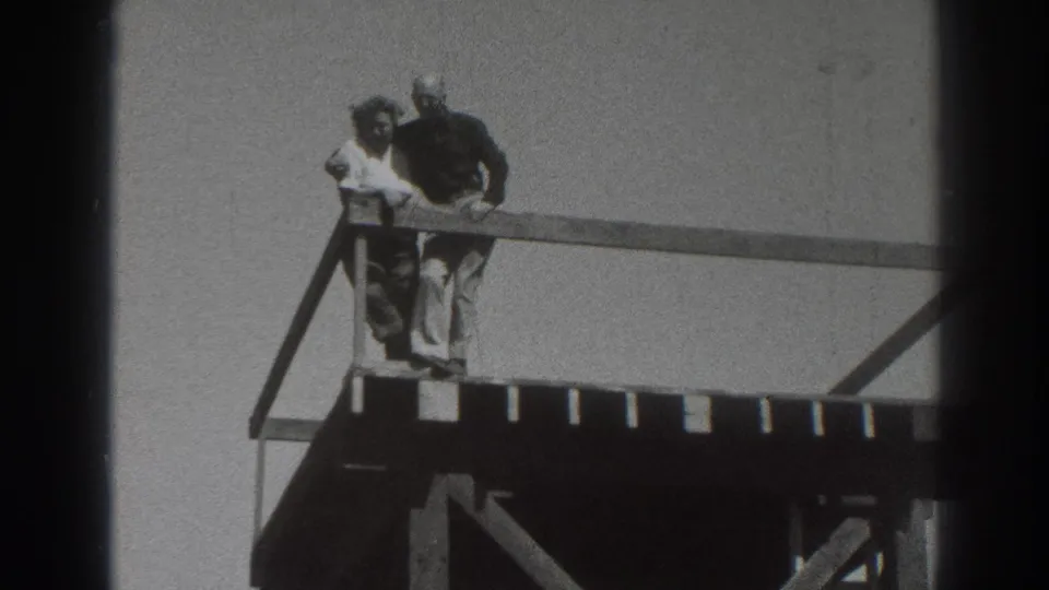 The couple remains at the platform railing, framed by wooden beams and open sky in black-and-white 8mm footage.