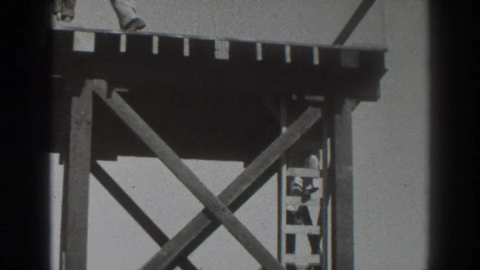 The 8mm frame shows the lower portion of the wooden scaffold, with the ladder and a partly visible worker framed by X-shaped braces.