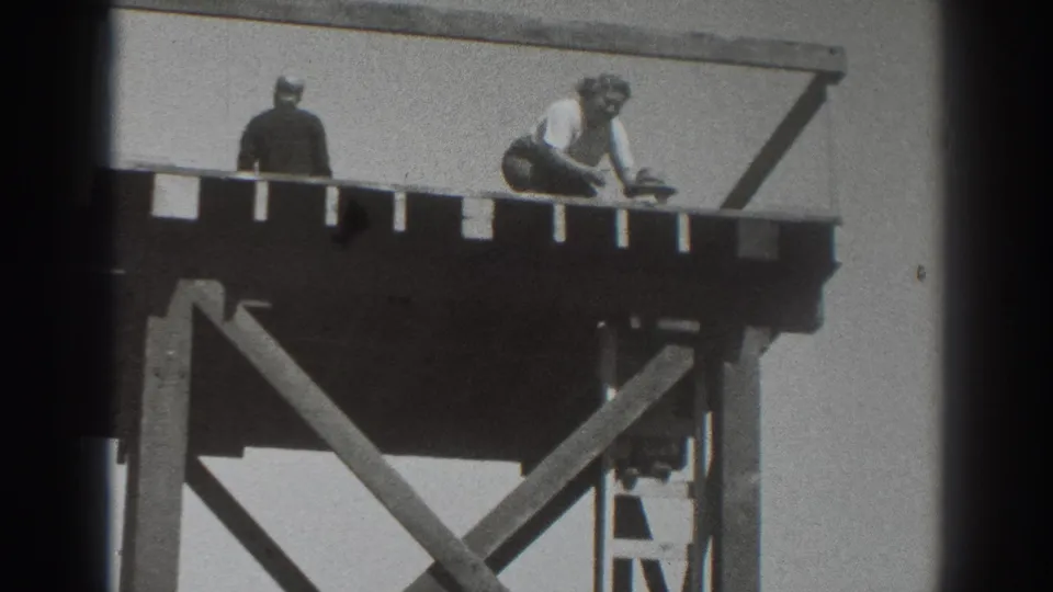 A person in light clothing crouches on the raised platform while another figure stands nearby in the 1937 home-movie footage.