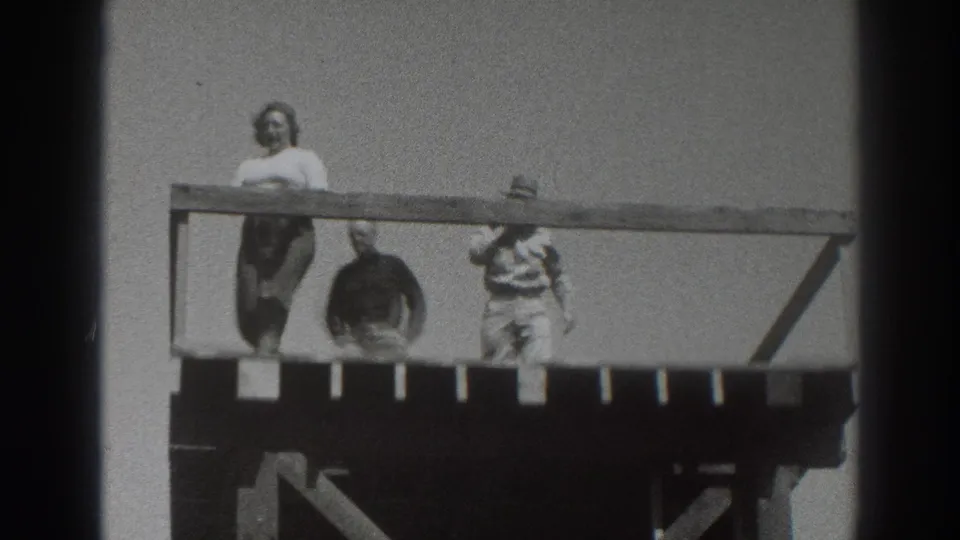 1937 Sacramento home-movie frame showing a woman in a light top and dark skirt near a hat-wearing man on the raised wooden deck.