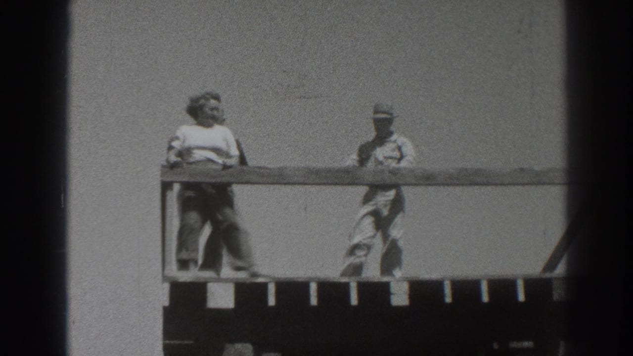 1937 Sacramento frame showing two adults moving along a wooden deck, with the railing crossing the foreground against a bright sky.