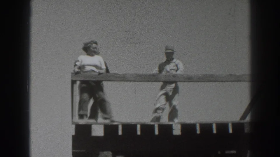 1937 Sacramento frame showing two adults moving along a wooden deck, with the railing crossing the foreground against a bright sky.