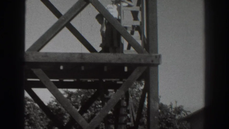 A person on a lower wooden platform near a ladder and cross-bracing, seen in black-and-white 8mm footage.