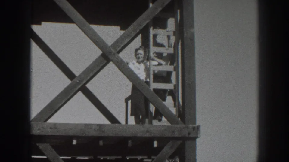 The woman remains on the scaffold platform, framed by diagonal bracing and a ladder against a bright sky.