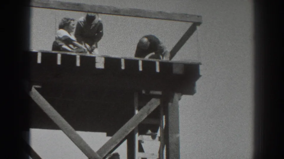 1937 Sacramento home-movie frame with two men working near the rail of an elevated timber platform and another figure partly obscured underneath.