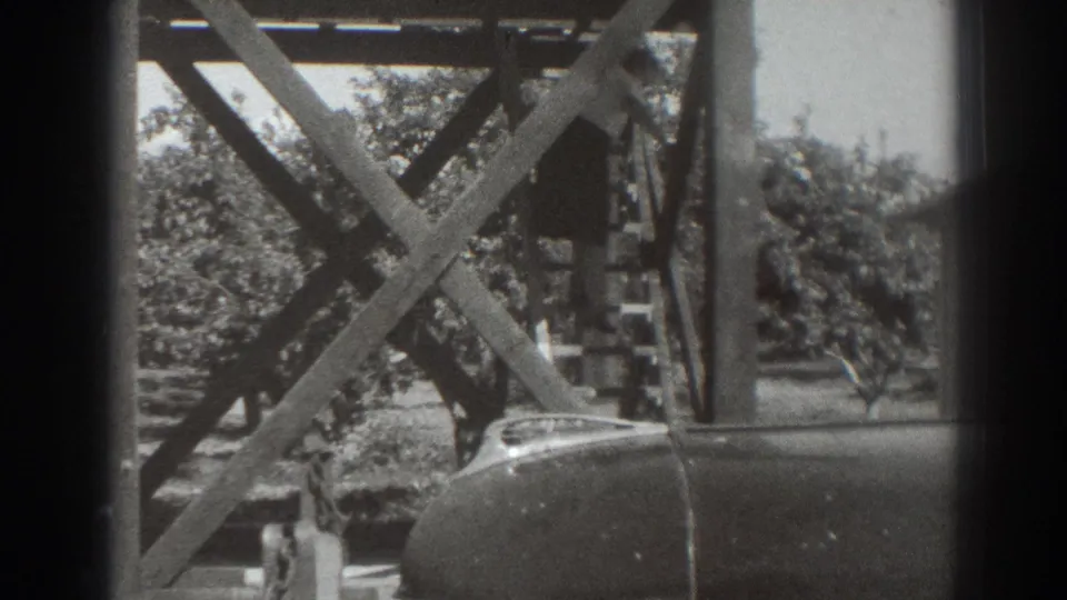 1937 Sacramento frame with crossed timber supports, leafy orchard trees, and a partial parked car in the foreground.