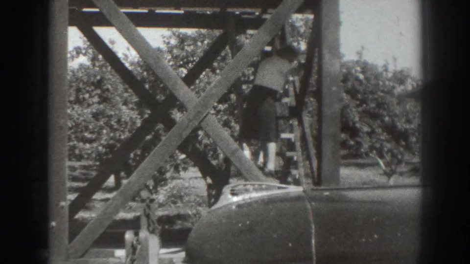 1937 Sacramento home-movie frame of a woman descending a steep exterior wooden stair near orchard trees and a parked car.