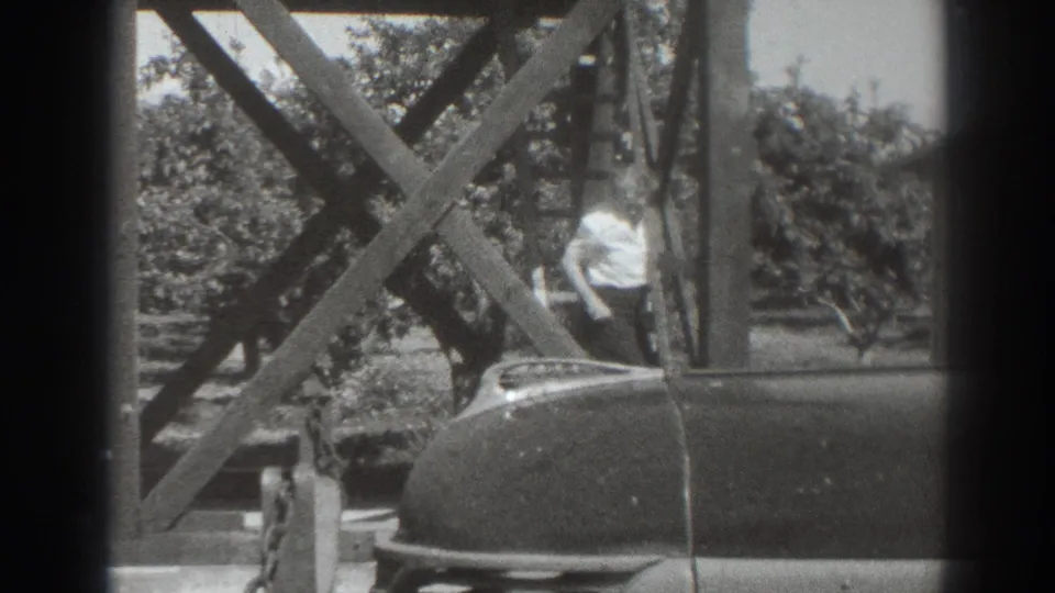 1937 Sacramento home-movie frame showing a woman near a car beneath a wooden structure, with trees in the background.