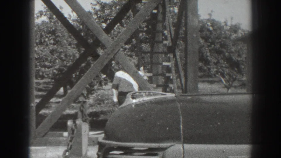1937 Sacramento home-movie frame with a woman leaning beside a parked car under wood bracing, with trees beyond.