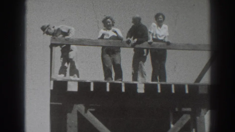 The left figure bends low beside the railing as three adults continue standing on the elevated wooden platform in Sacramento home-movie footage.