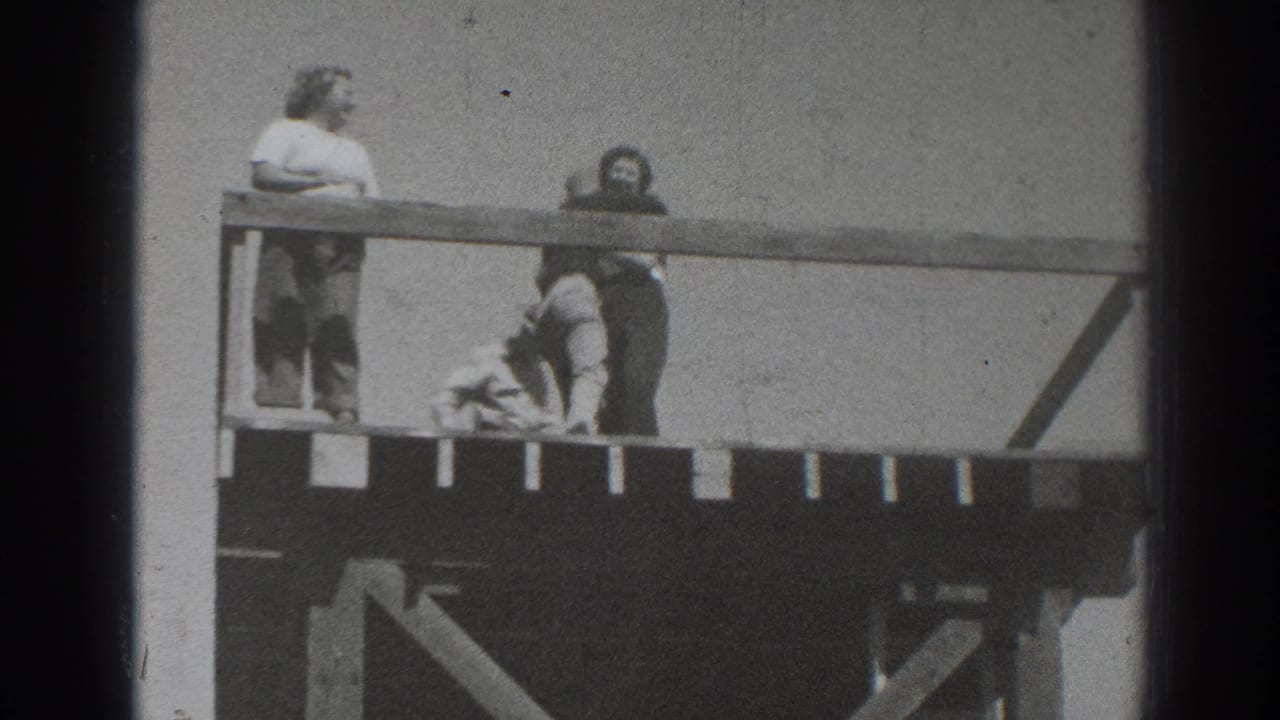 Two people stand on or beside an elevated wooden structure in a grainy black-and-white frame.