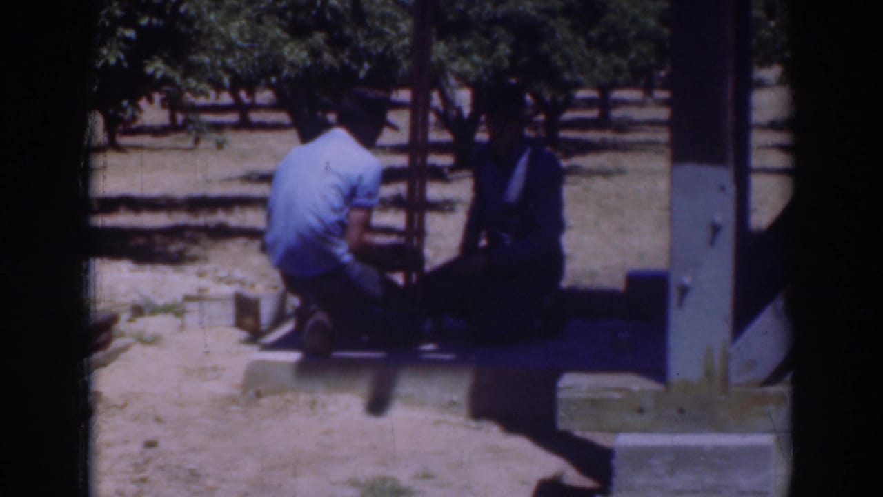 1937 Sacramento frame showing a man in a light shirt kneeling beside another man in darker clothing near a wooden frame outdoors.