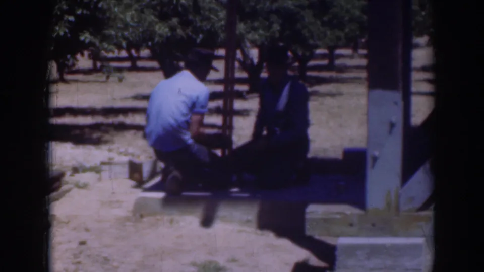 1937 Sacramento frame showing a man in a light shirt kneeling beside another man in darker clothing near a wooden frame outdoors.