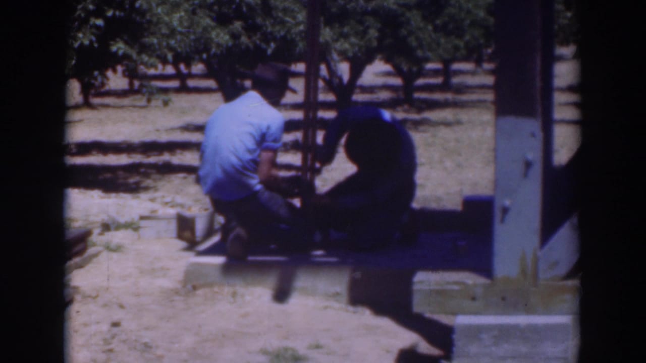 1937 Sacramento home-movie frame with two men bent over a wooden platform or frame, surrounded by trees and dry ground.