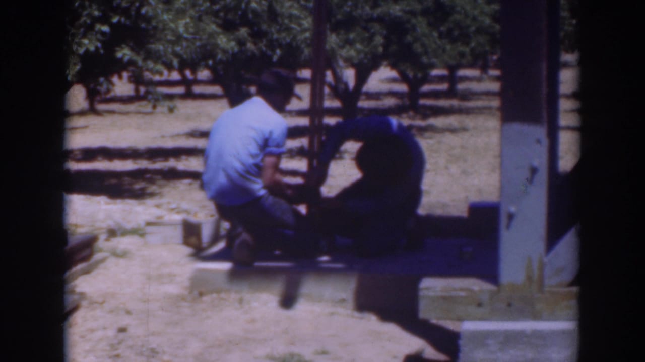 1937 Sacramento frame showing two men crouched near a wooden support, with orchard-like trees, dry soil, and strong midday shadows.