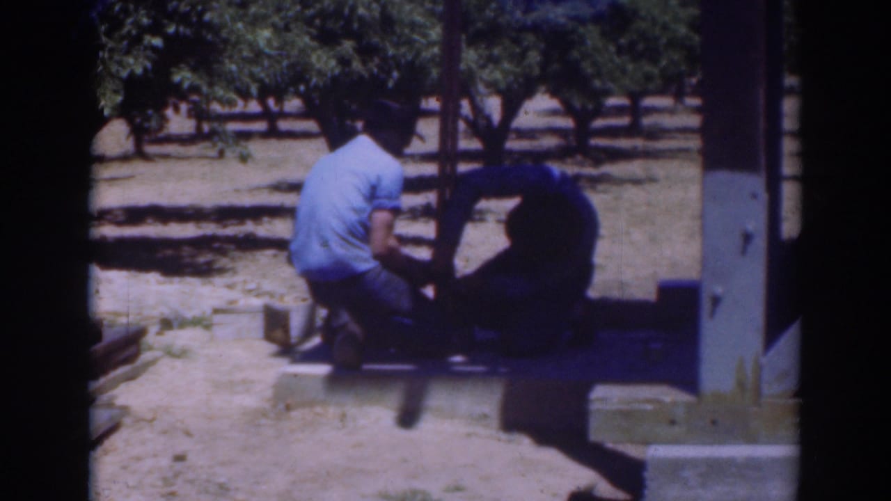 Two men crouch by a shaded structure while handling a post outdoors.