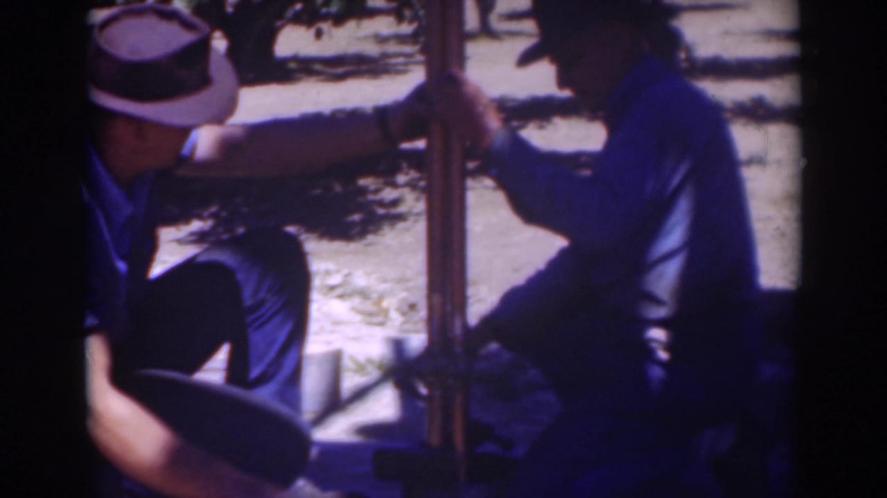 Closer 8mm frame of two middle-aged men in hats working with a vertical post outdoors in the Sacramento, California clip.