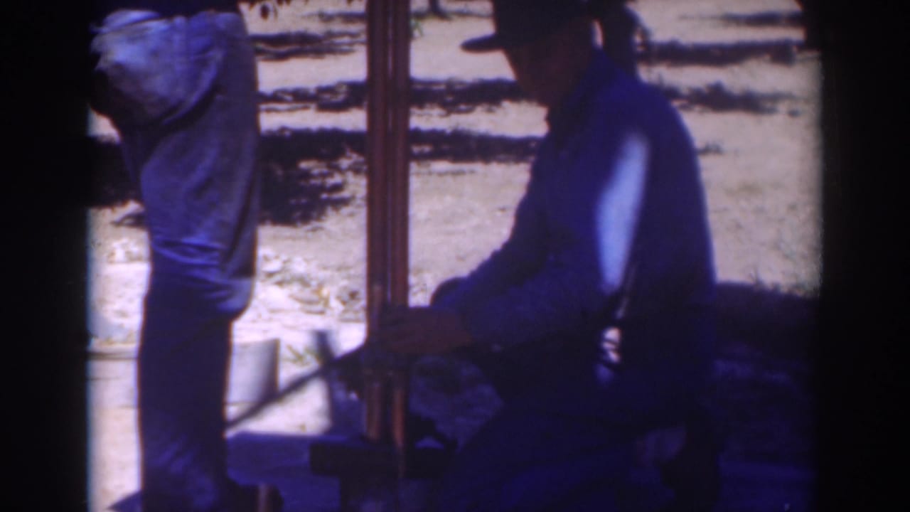 Two men outdoors beside a vertical pole or pipe, one kneeling near its base.