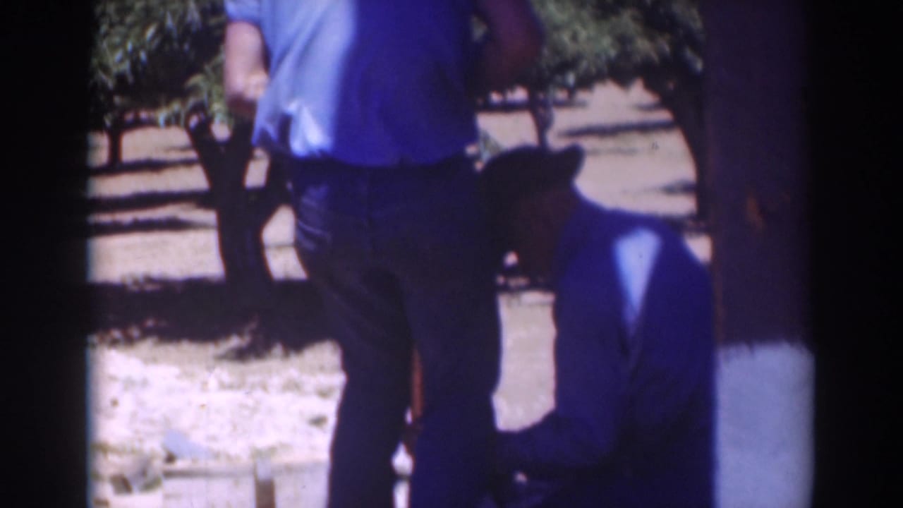 Two men outdoors near trees, one standing and one kneeling beside a wooden post or work area.