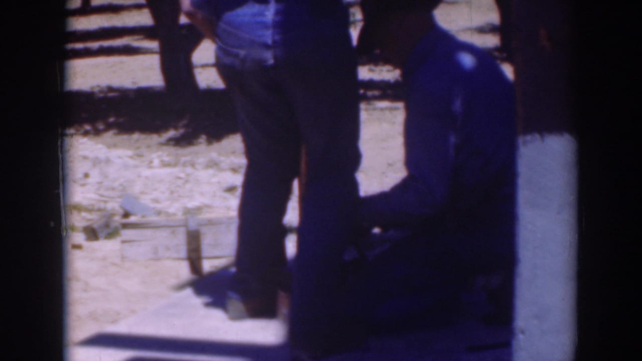 A standing man and a seated or kneeling man work beside a wooden structure outdoors.