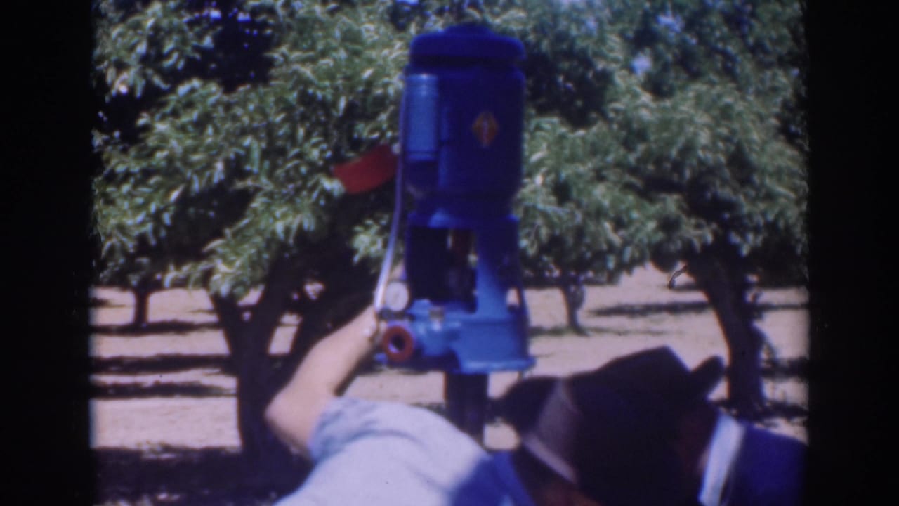 The blue pump is centered against orchard rows while a worker below reaches toward its lower fittings.