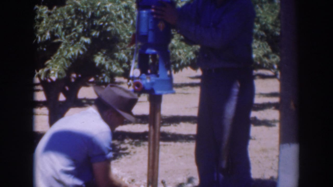 Two adult men in work clothes stand and crouch around the blue pump in the Sacramento orchard, continuing the repair.