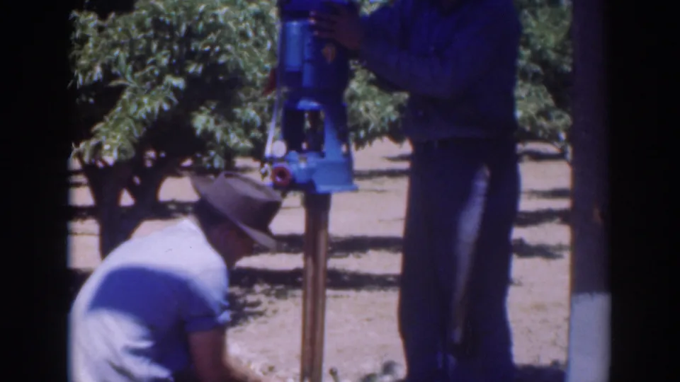 Two adult men in work clothes stand and crouch around the blue pump in the Sacramento orchard, continuing the repair.