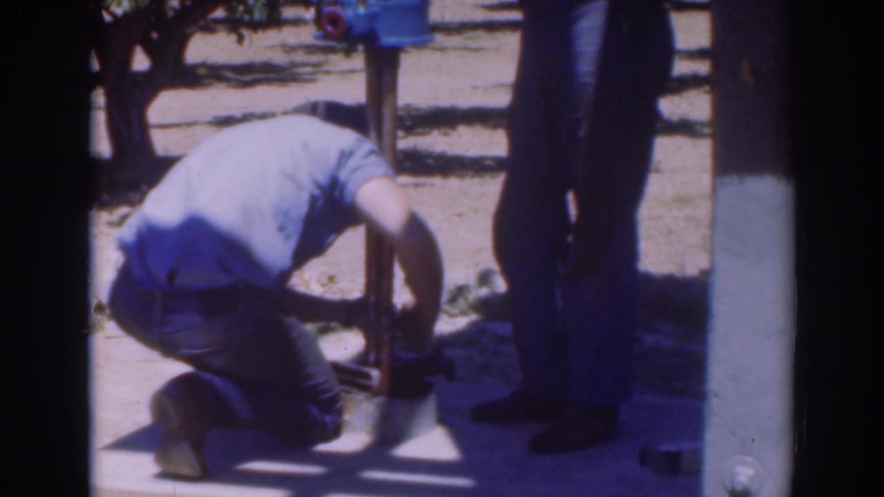 Man kneels beside a blue pump while another man stands nearby outdoors.
