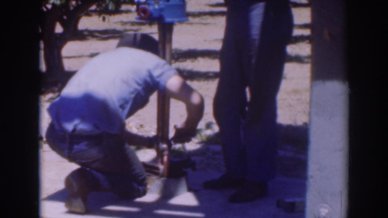 The kneeling worker remains focused on the lower pump hardware, with another adult partly visible beside the equipment outdoors.