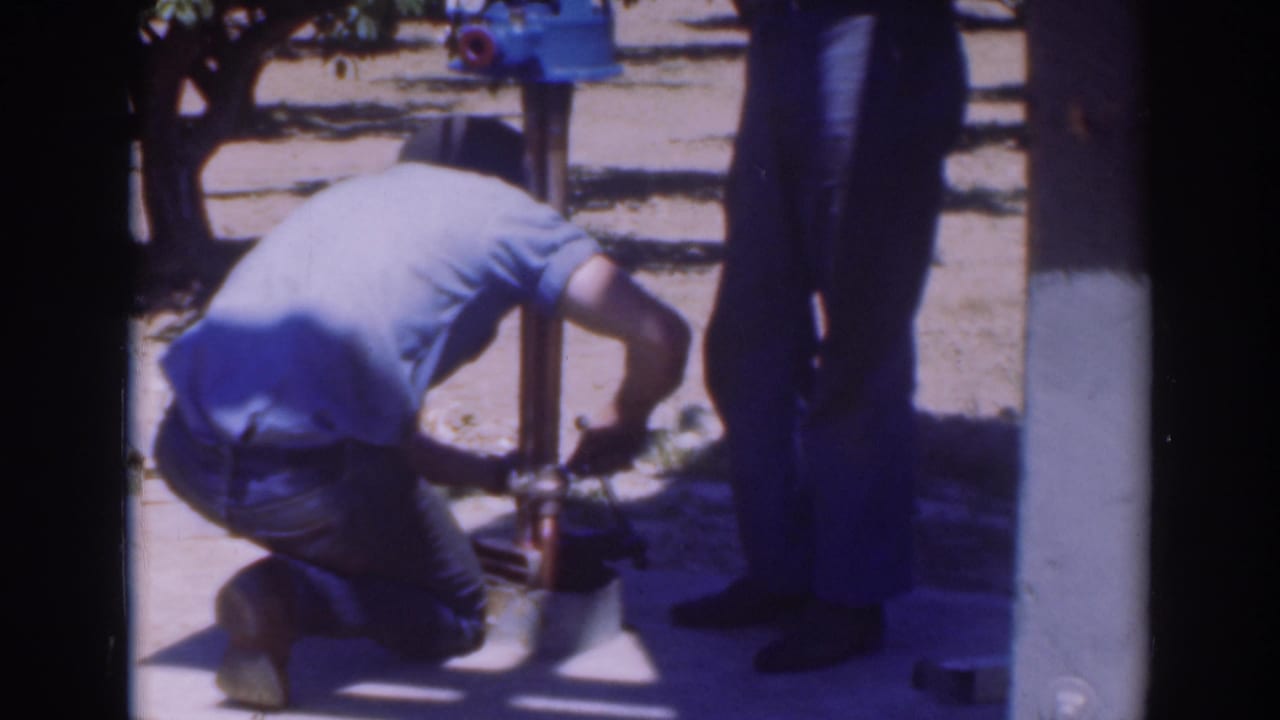 A crouching man in a light shirt works low on the pump assembly, with another person standing at the edge of the 8mm frame.