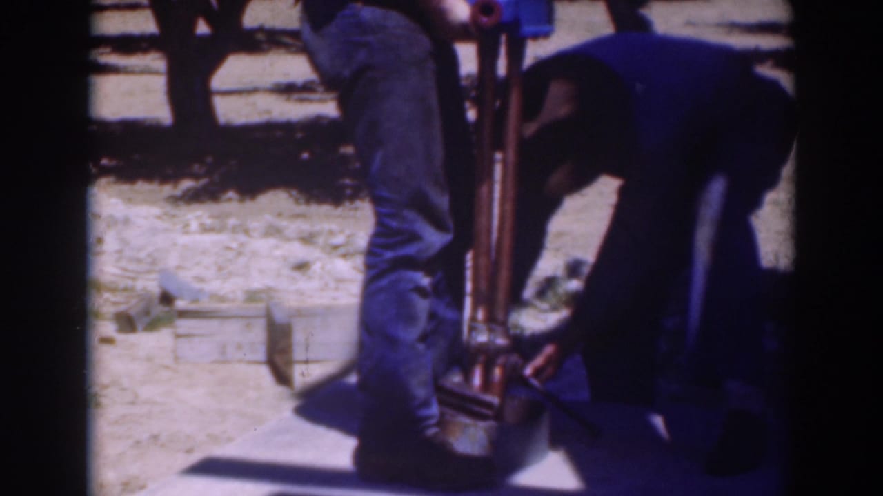 1937 home-movie frame from Sacramento showing two men focused on the lower part of a large tool in an orchard.