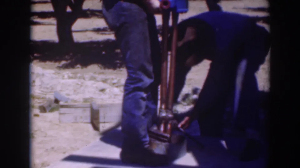1937 home-movie frame from Sacramento showing two men focused on the lower part of a large tool in an orchard.