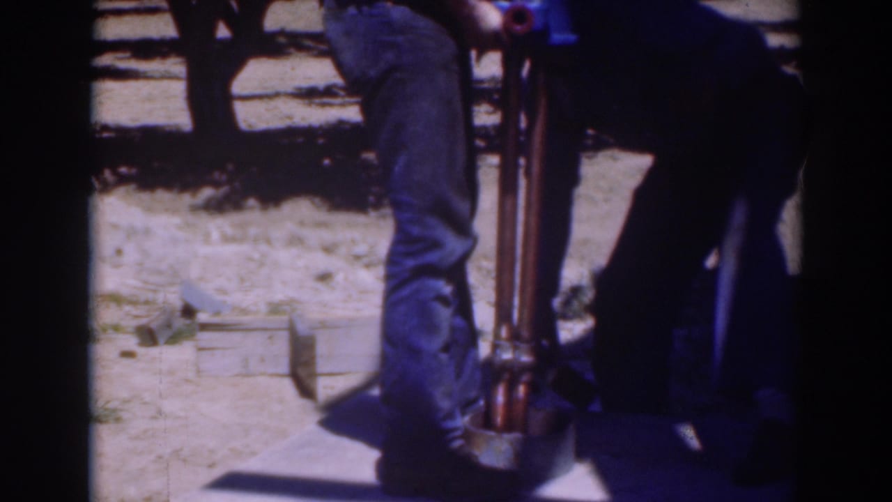 Two adults in jeans stand over a vertical tool on dry ground in bright sun.