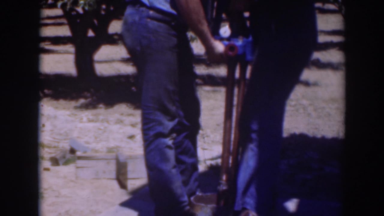 Two adult men continue working around a blue ground object and upright tool in a sunny Sacramento outdoor setting.