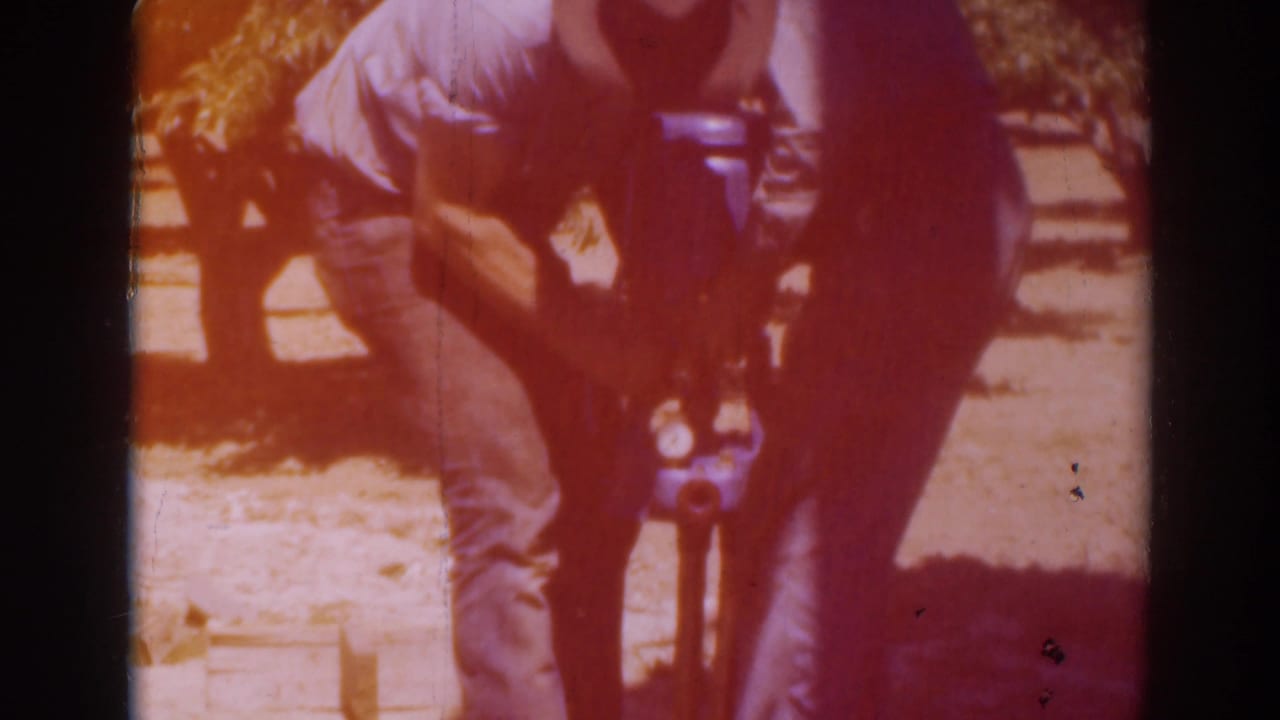 Two men bend over the upright tool and blue ground object as warm exposure flare colors the 1937 Sacramento 8mm frame.