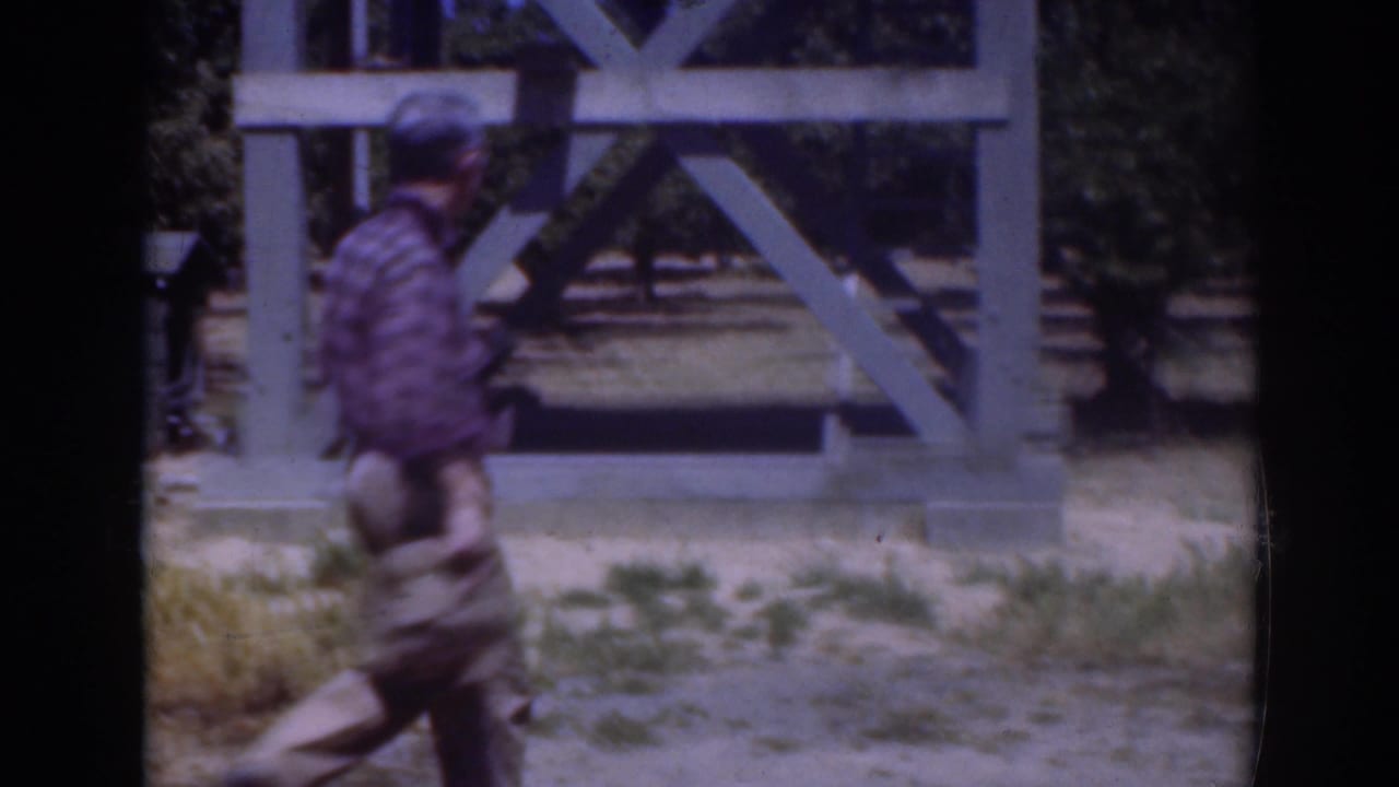 In 1937 Sacramento footage, a man in a blue plaid shirt bends near grass in front of a large pale wooden frame.