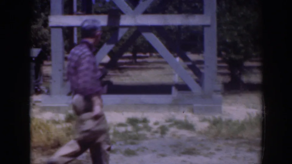 In 1937 Sacramento footage, a man in a blue plaid shirt bends near grass in front of a large pale wooden frame.