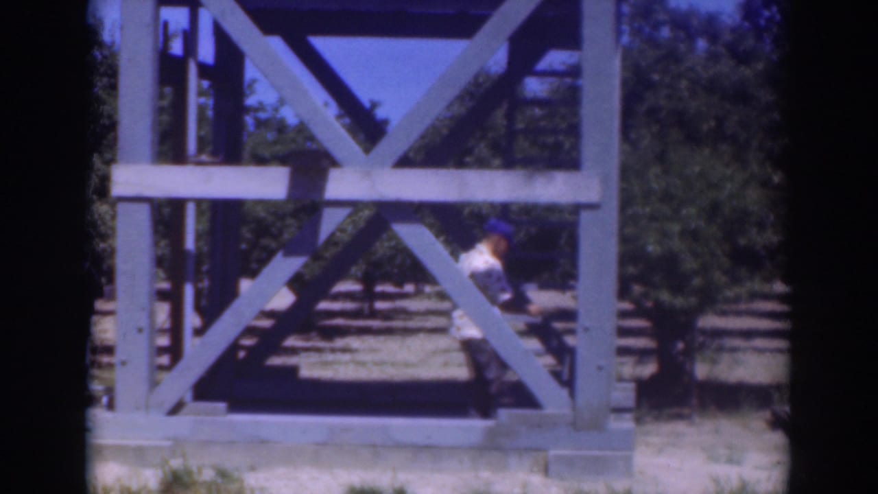 1937 Sacramento frame showing a man in a blue cap near the interior stairs of a painted wooden tower, with trees and dry ground behind.