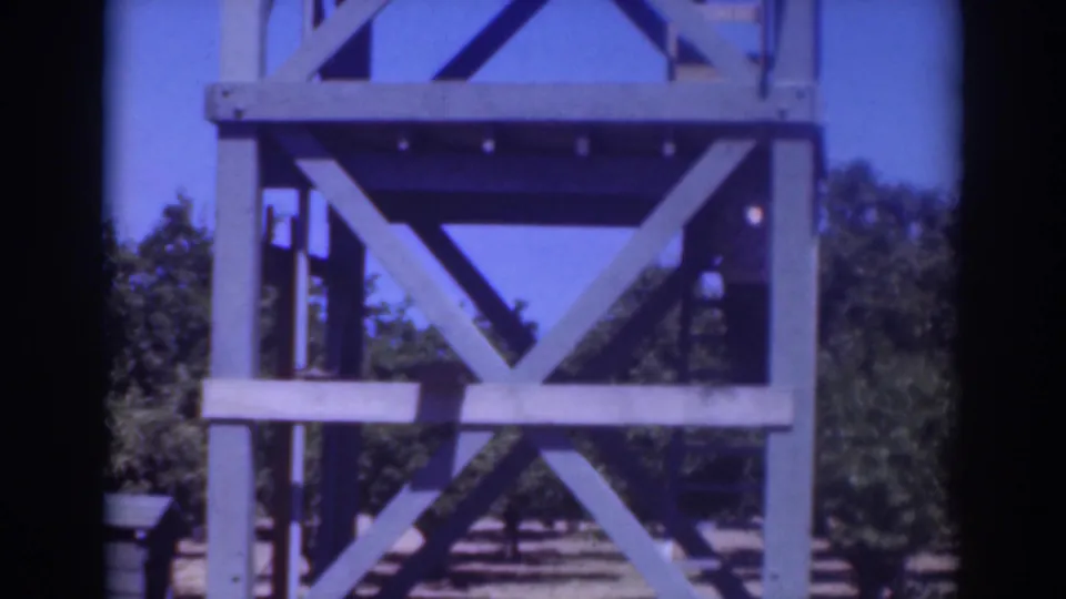 1937 Sacramento home-movie frame showing the blue wooden tower from closer range, with trees and dry ground beyond.