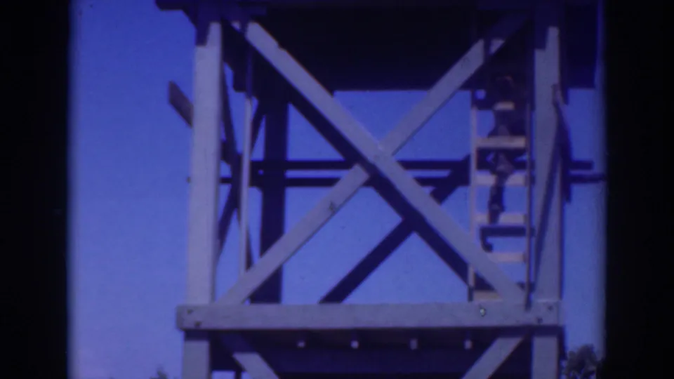 1937 Sacramento 8mm frame of a person climbing along the right side of a tall metal tower, with bracing silhouetted against blue sky.