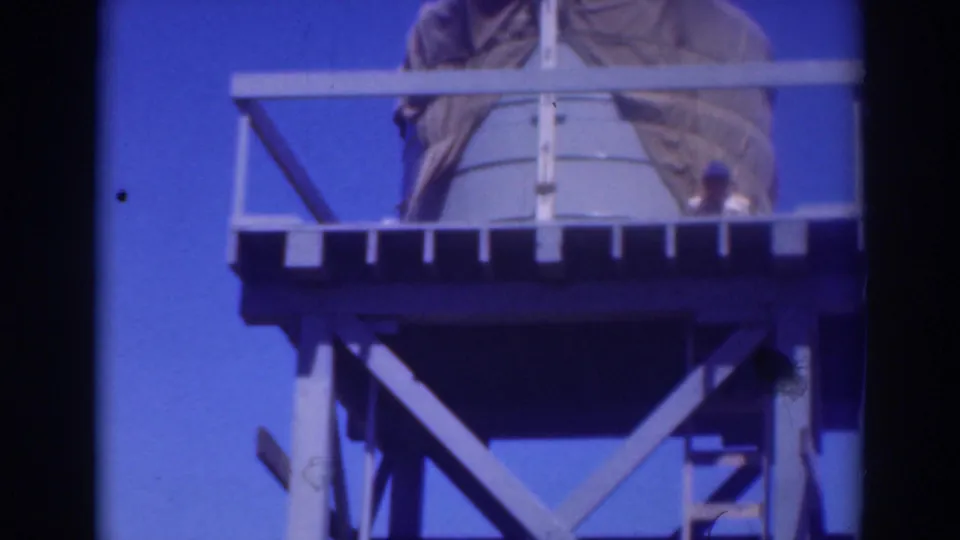 A low-angle frame shows the Sacramento tower's platform, ladder, elevated tank, and dark bracing, with a small worker figure partly visible near the rail.