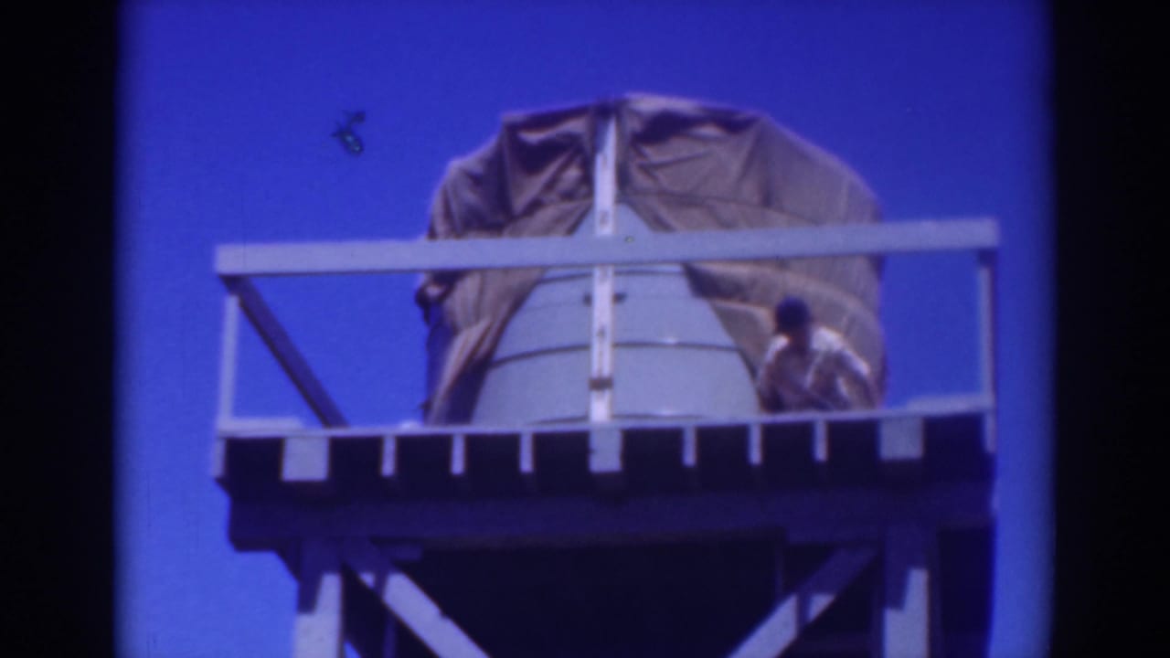 Worker crouches near the right side of a tarp-covered tank on a tower platform.