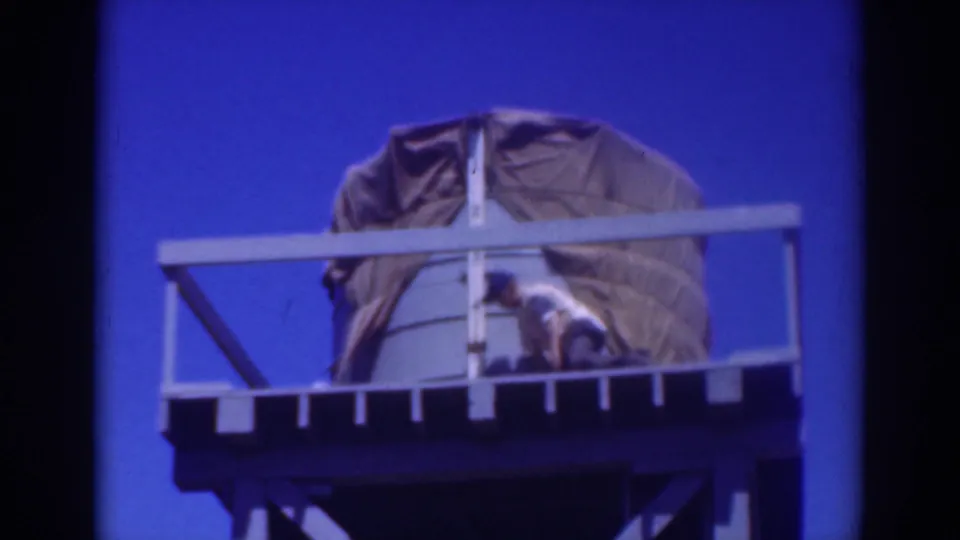 The worker bends low on the elevated water tower platform, appearing to adjust the tarp around the tank.