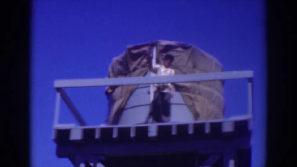 A low-angle 8mm frame shows the person on the high platform in Sacramento, with railings cutting across the body and a covered object behind.